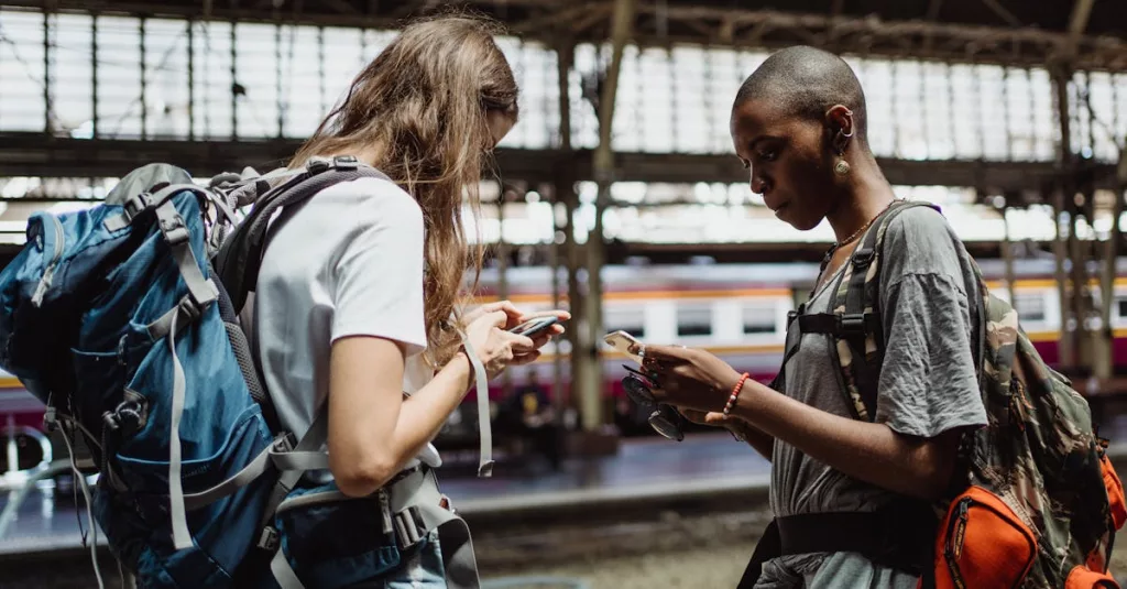 Two adventurous backpackers using smartphones while waiting at a bustling train station.